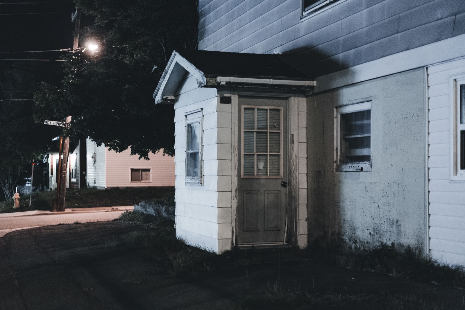 An aging painted wood entry door with a nine-lite glass panel in a small clapboard entry vestibule on a white wood-sided American house, lit by a street lamp at night