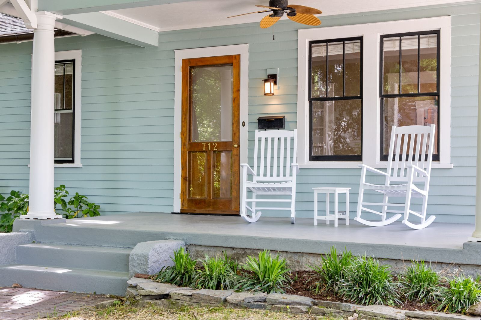 Full-view glass storm door over a natural wood entry door on the covered porch of a light-blue bungalow with two white rocking chairs
