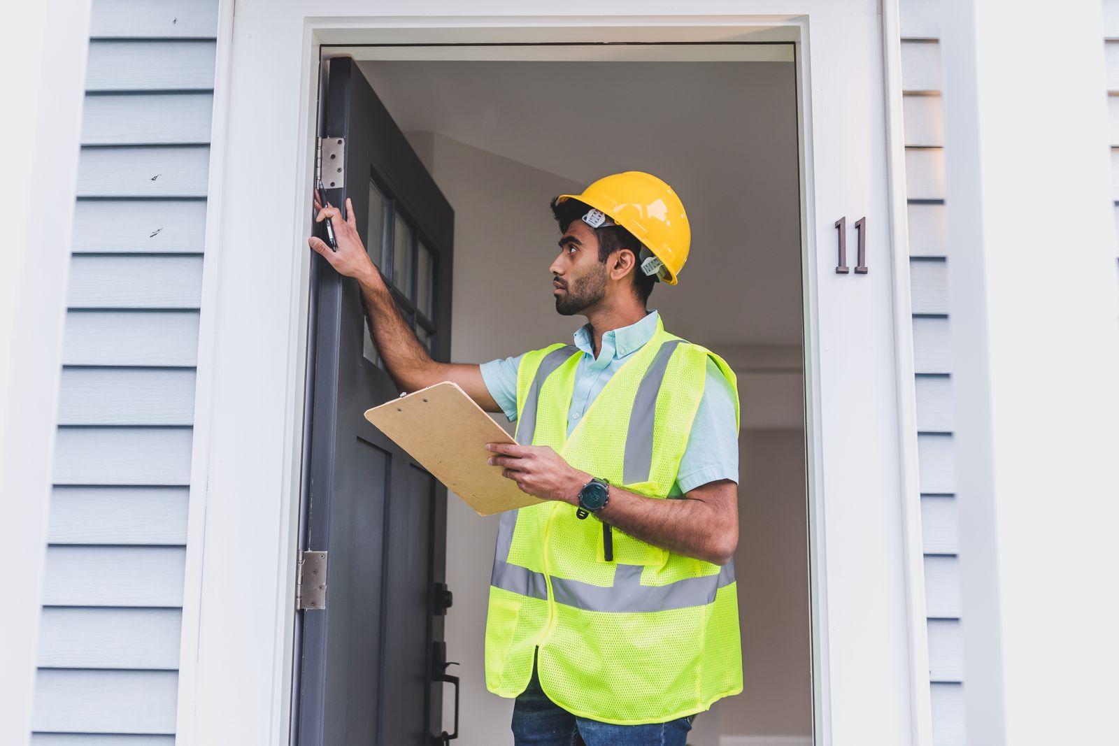 An installer in a hard hat and hi-vis vest standing in the open front doorway of a pale grey lap-siding house, one hand on the hinge side of a dark entry door, checking the upper hinge