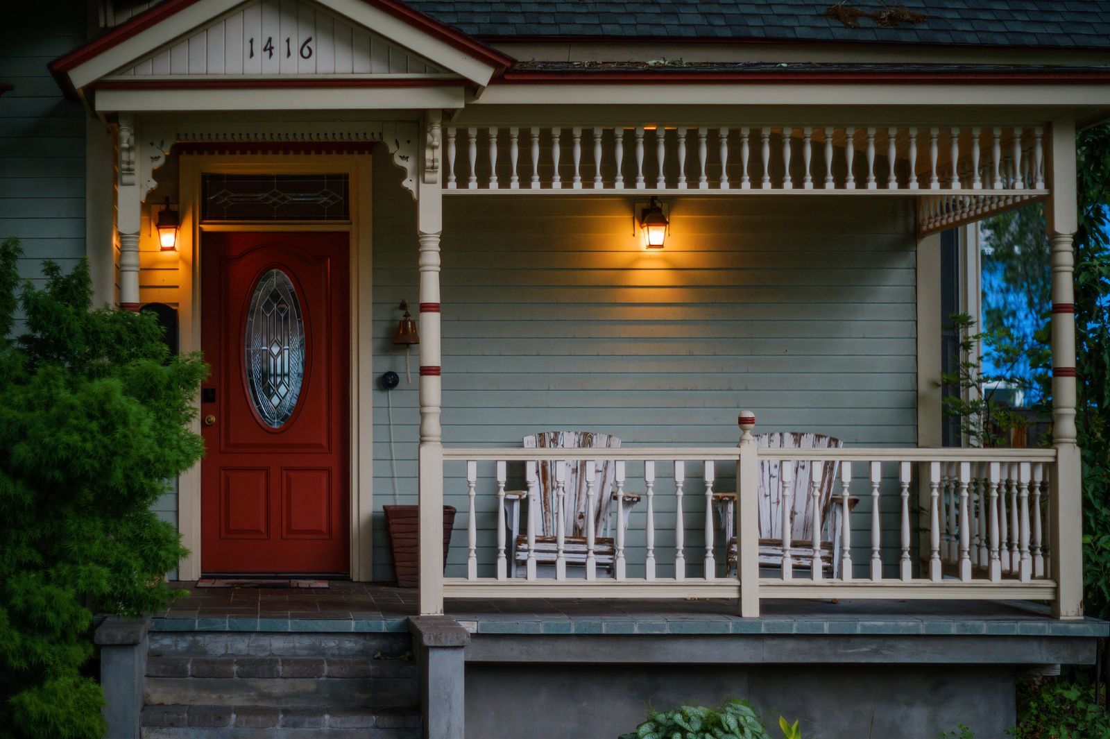Red entry door with an oval glass lite on a covered front porch with wooden armchairs and porch lights at dusk