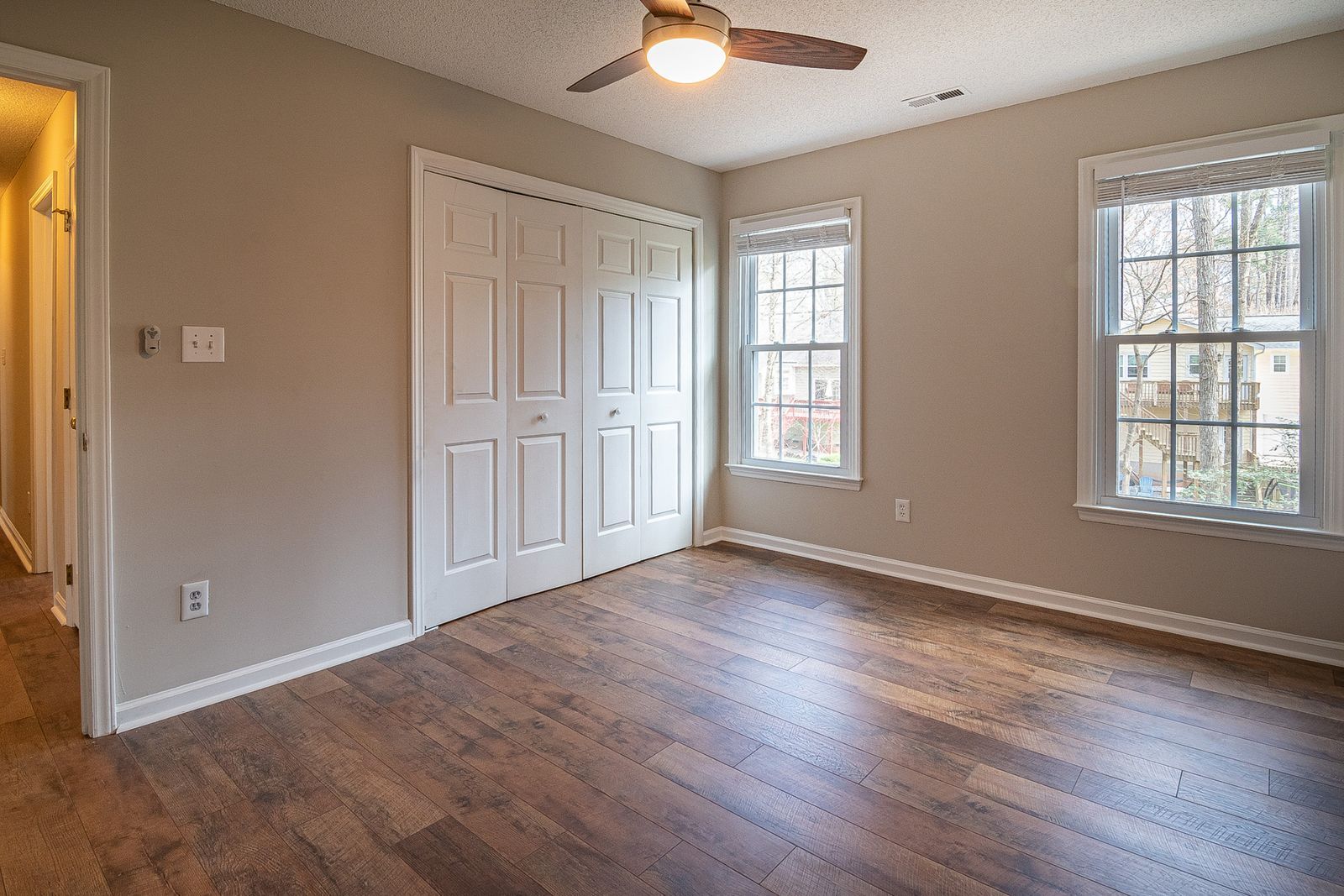 Empty bedroom in an American home with a white six-panel bi-fold closet door, white casing and baseboard, a second white panel door at the room entry, double-hung grilled windows and a ceiling fan